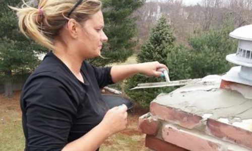 A woman applies mortar to the top of a brick chimney with a trowel outdoors on a cloudy day.